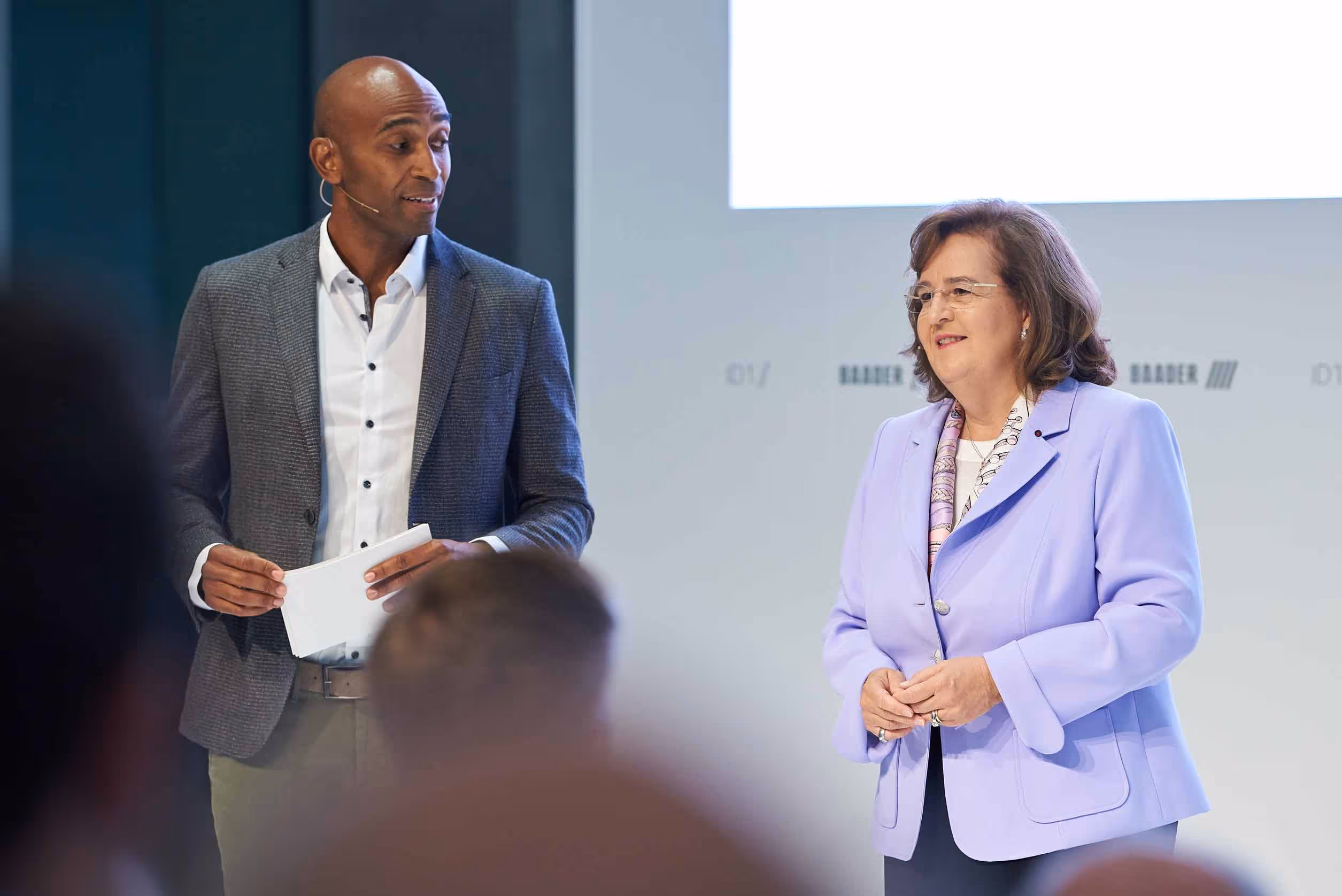 Two professionals speaking on stage during a conference, a man holding notes and a woman wearing a lavender blazer with a patterned scarf.