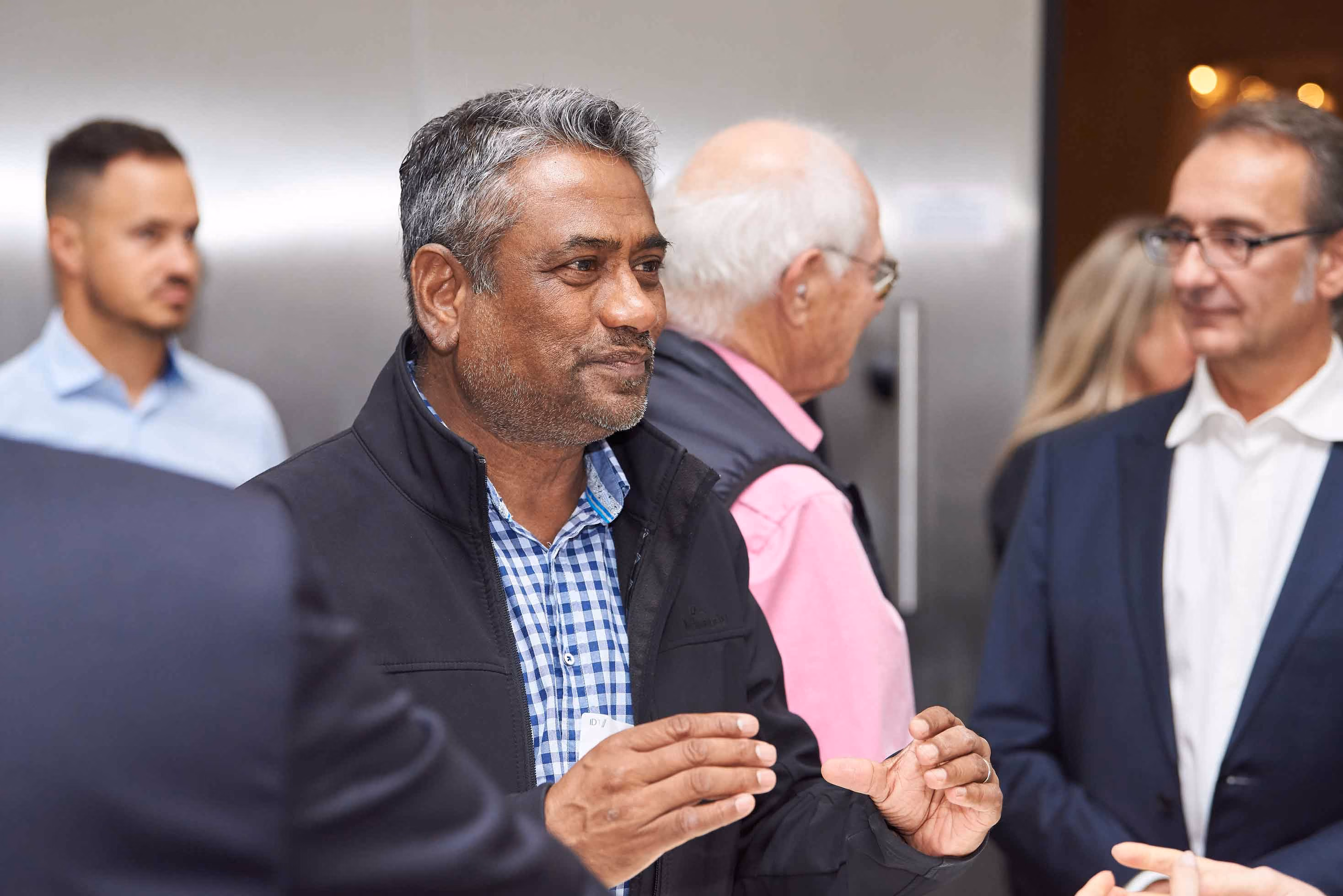 Man in a black jacket and blue checkered shirt talking and gesturing with his hands in a social gathering with several people.