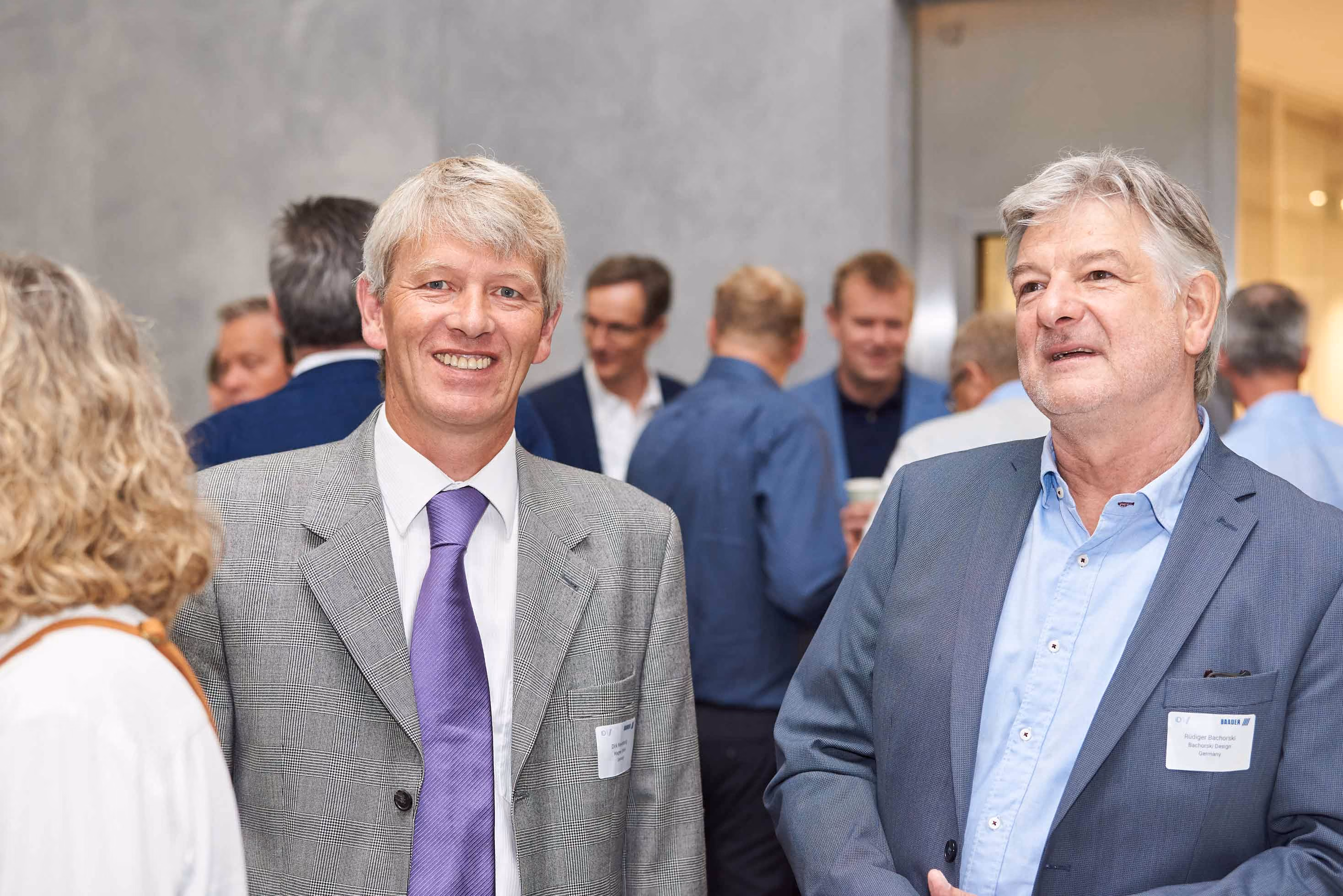 Two middle-aged men in suits at a professional networking event with other attendees in the background.