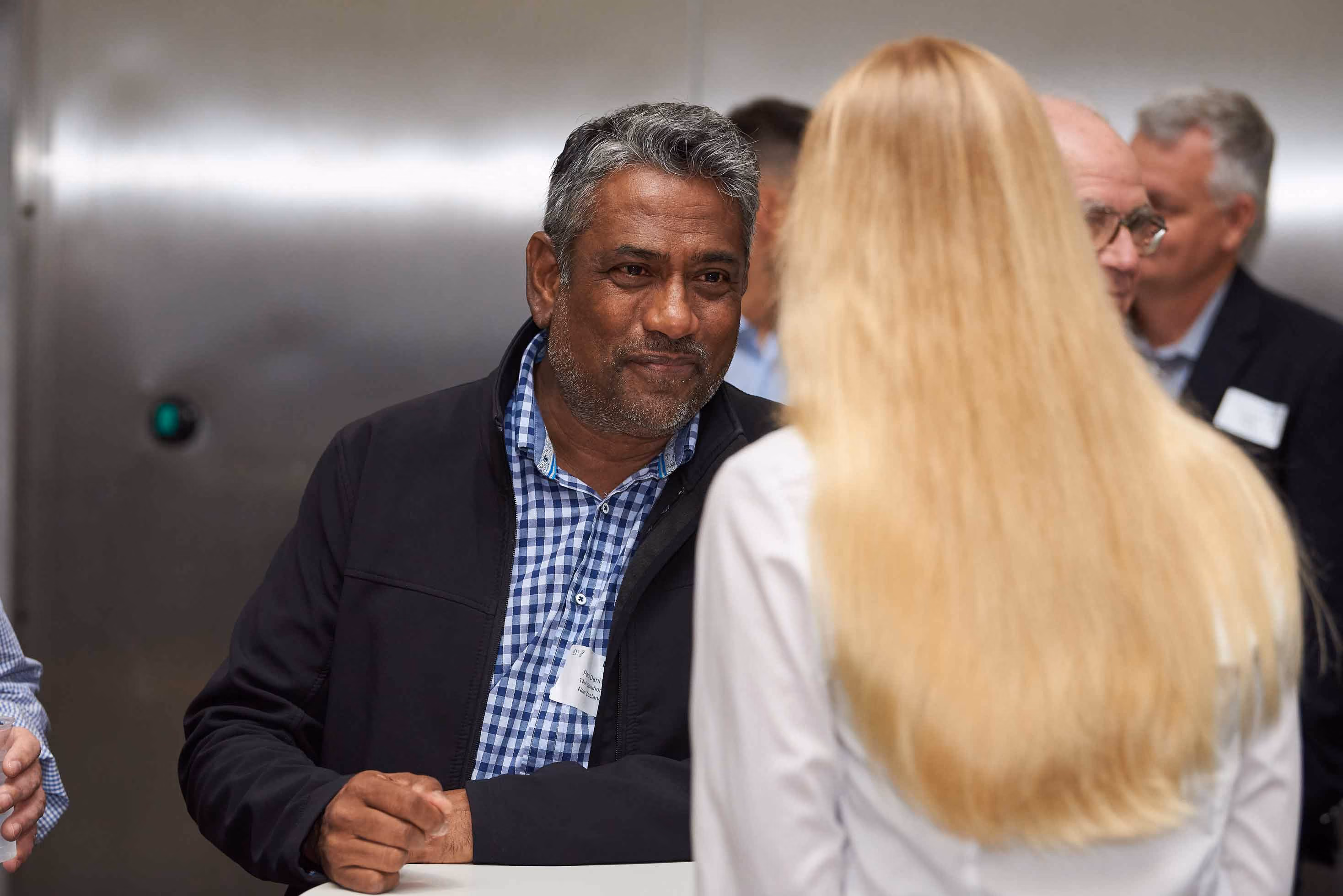 Man in a checkered shirt and jacket talking to a woman with long blonde hair at an event.