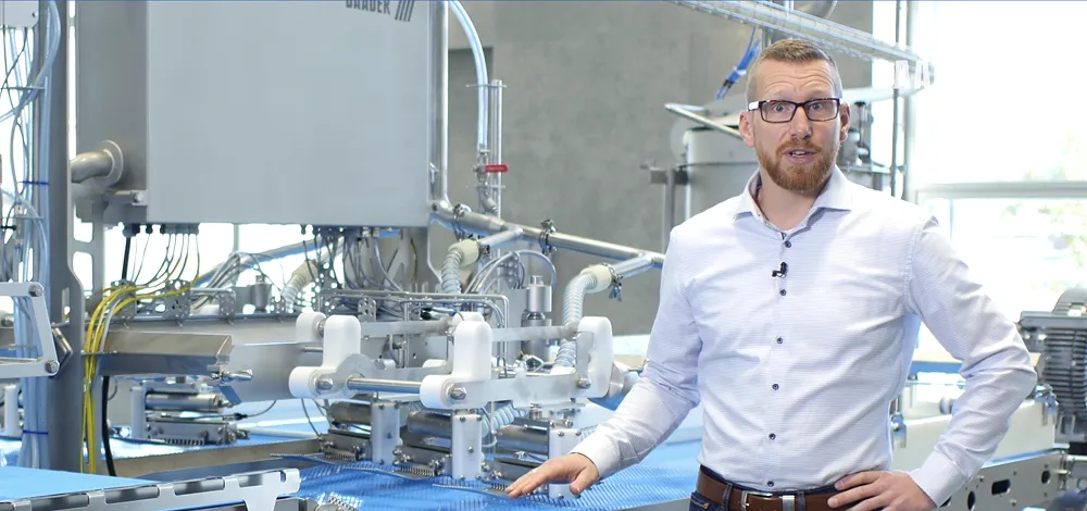Man wearing glasses and a light blue shirt standing next to industrial machinery in a factory setting.
