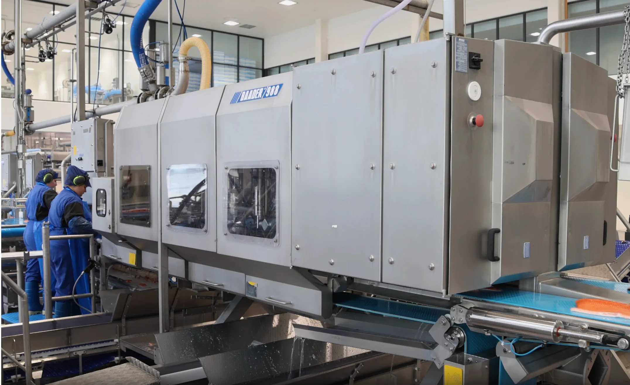 Industrial fish processing machine labeled Baader 988 with workers in blue uniforms and hearing protection operating it.