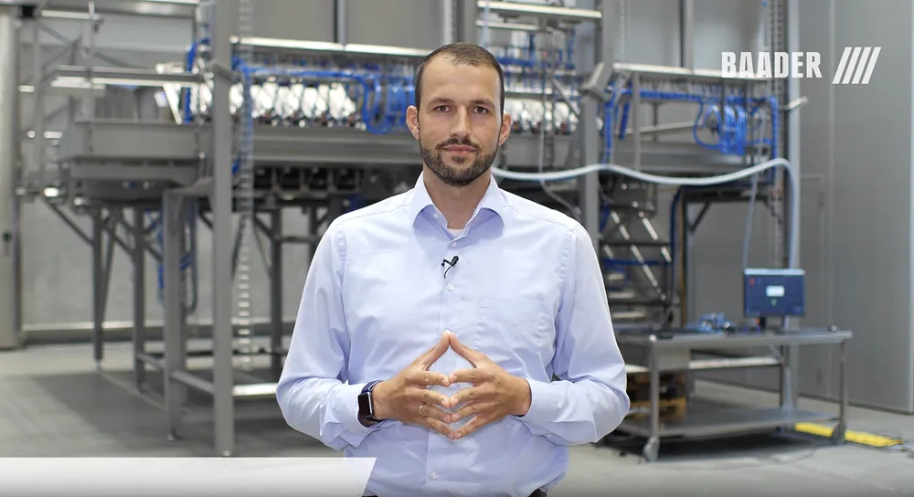 Man in light blue shirt standing with hands clasped in front of industrial machinery inside a factory.