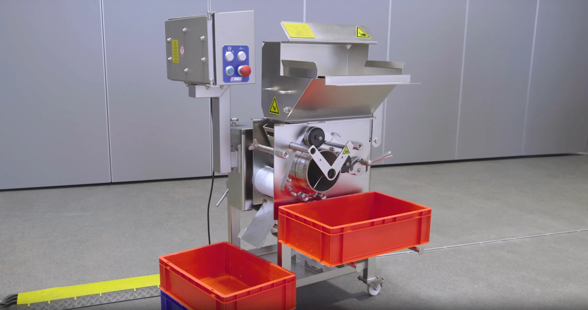 Industrial stainless steel machine with control panel and two orange plastic crates, placed on a gray floor.