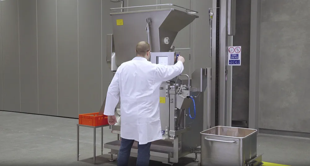 Man in white lab coat operating industrial food processing machine with stainless steel container nearby.