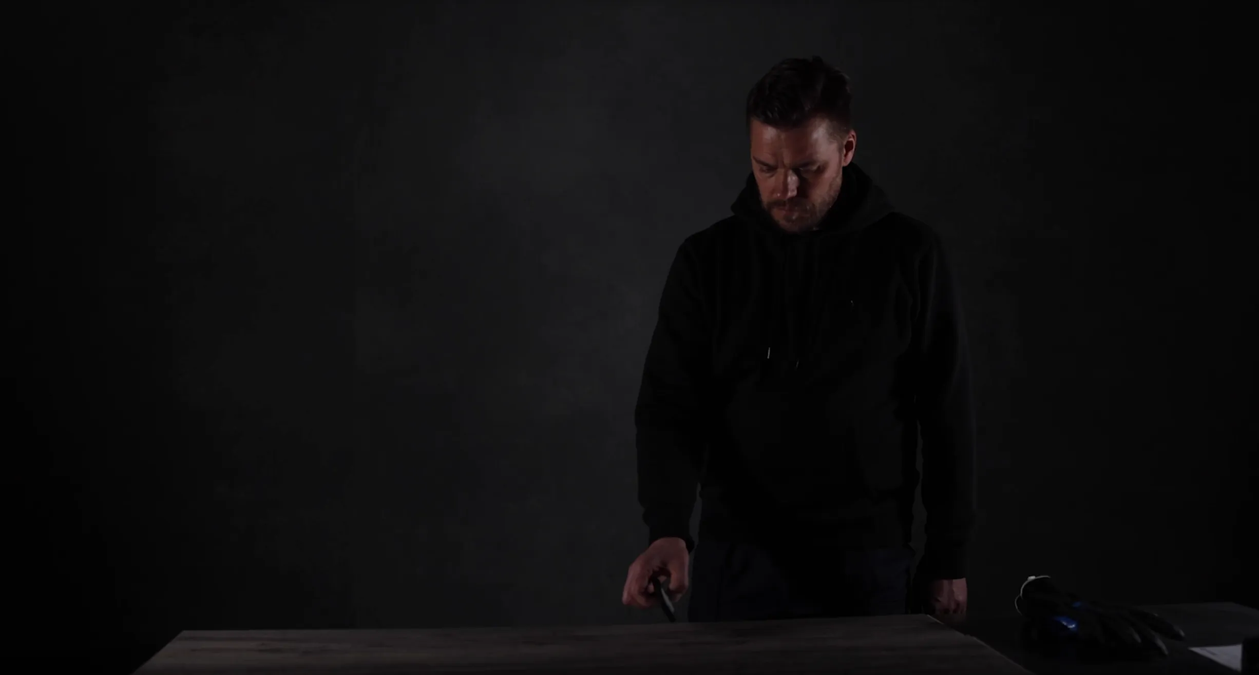 Man in a black hoodie looking down and holding an object over a wooden table with gloves on the side.