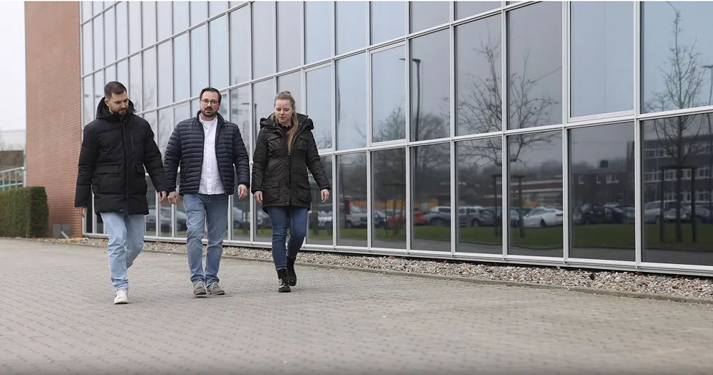Three people wearing winter jackets walking together outside near a large glass building on a cloudy day.
