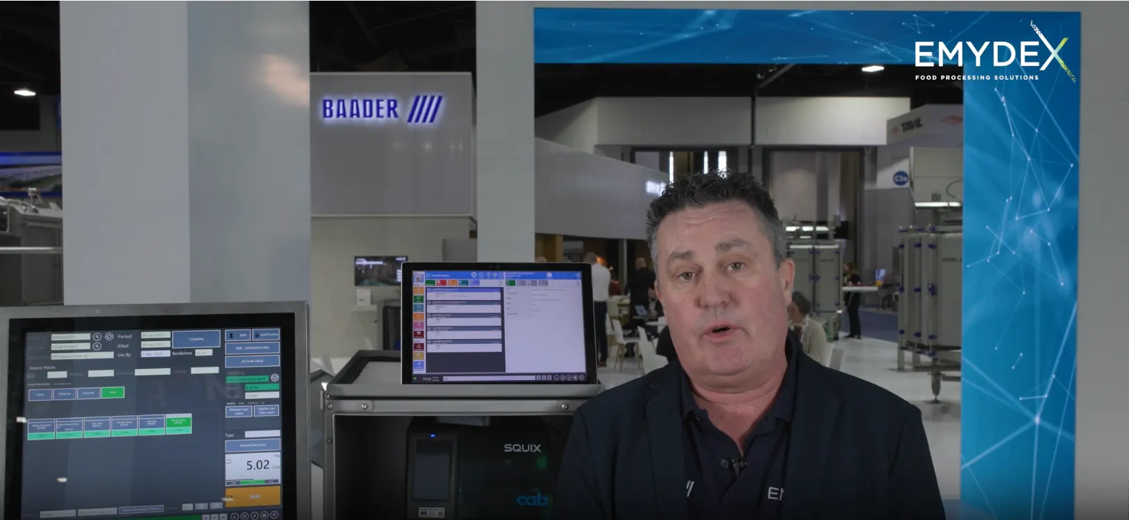 Man speaking in a trade show booth with computer screens displaying industrial software and equipment behind him.
