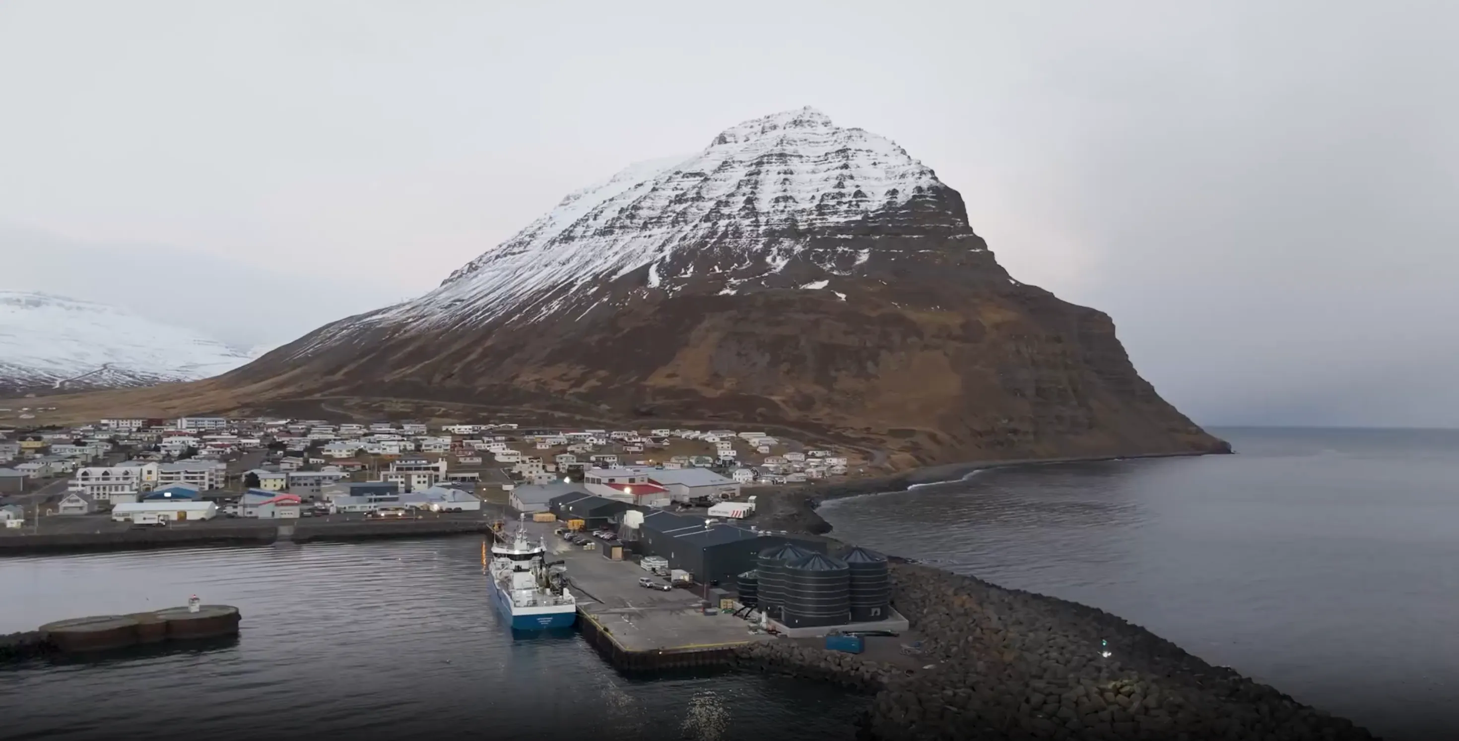 Small coastal town with a docked fishing boat beside a pier and large silos, set against a steep mountain partially covered in snow.