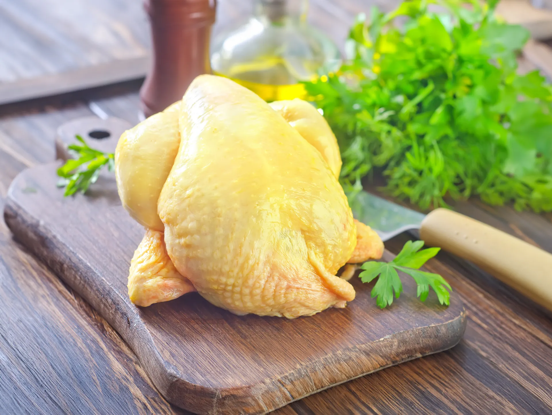 Whole raw yellow chicken placed on a wooden cutting board with fresh green herbs and a knife in the background.