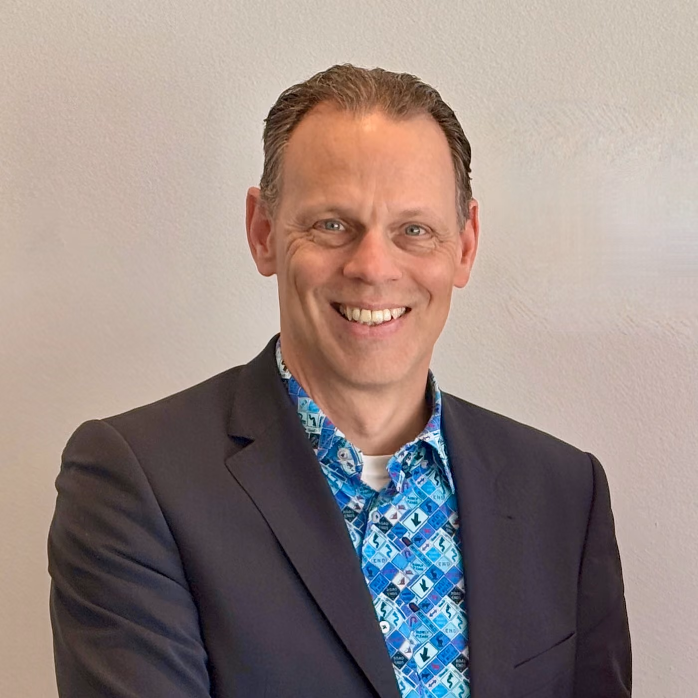 Smiling man in a dark blazer and blue patterned shirt standing against a plain light wall.