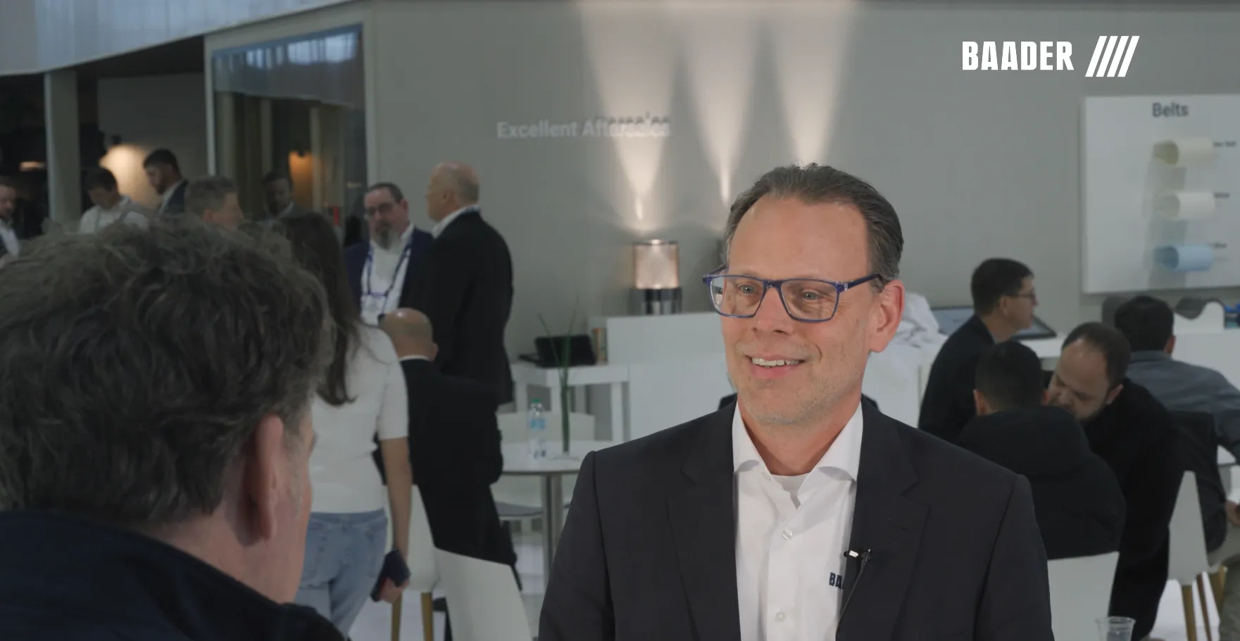 Man in glasses and business attire smiling and talking to another person in a busy, modern conference or trade show setting.