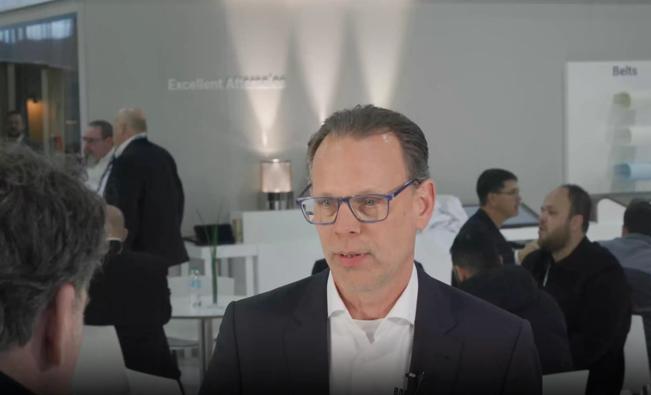 Man wearing glasses and a dark suit speaking with another person in a professional setting with people in the background.