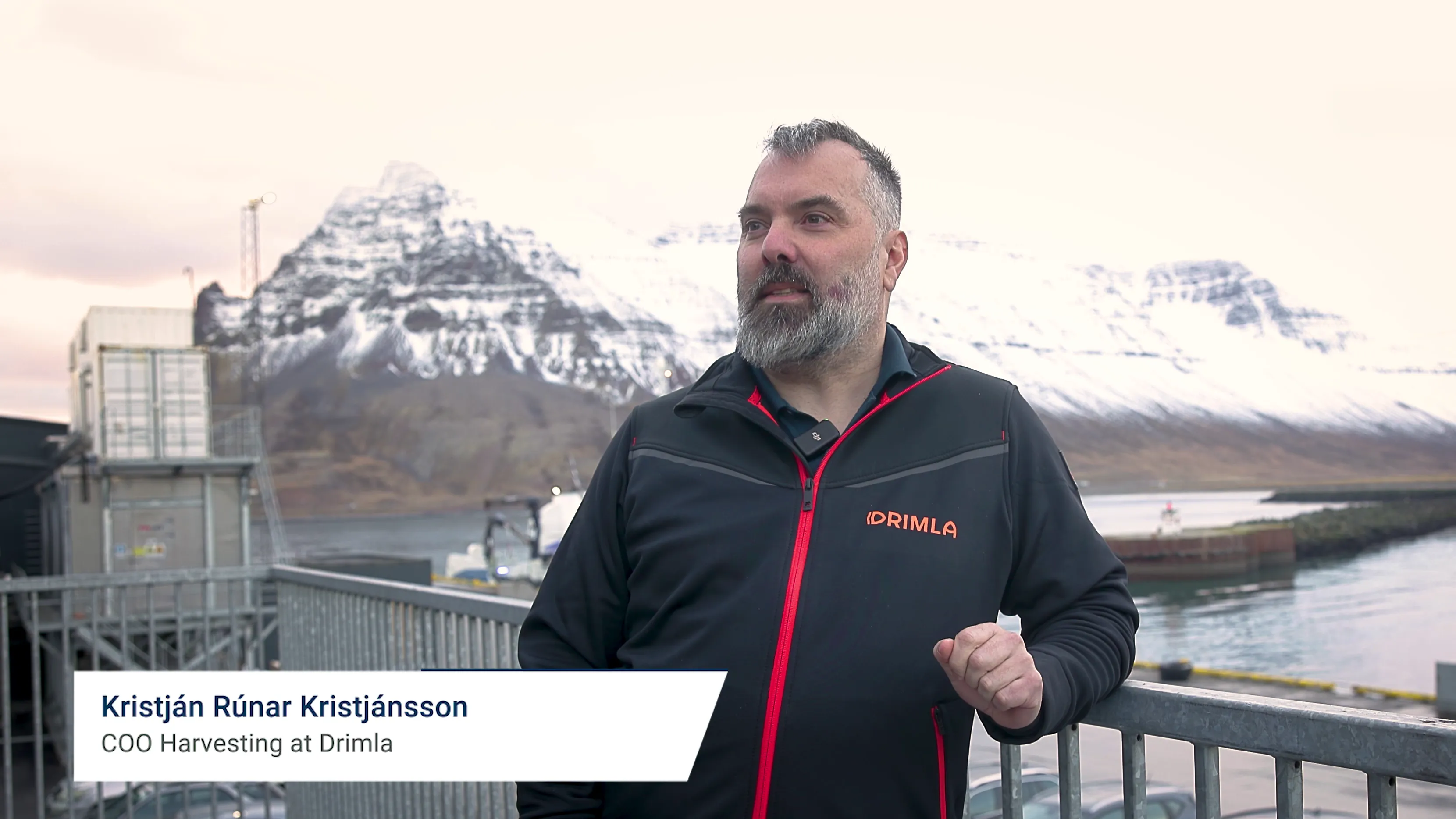 Man wearing a black jacket with red zipper and Drimla logo standing by a metal railing with snowy mountains and a harbor in the background.