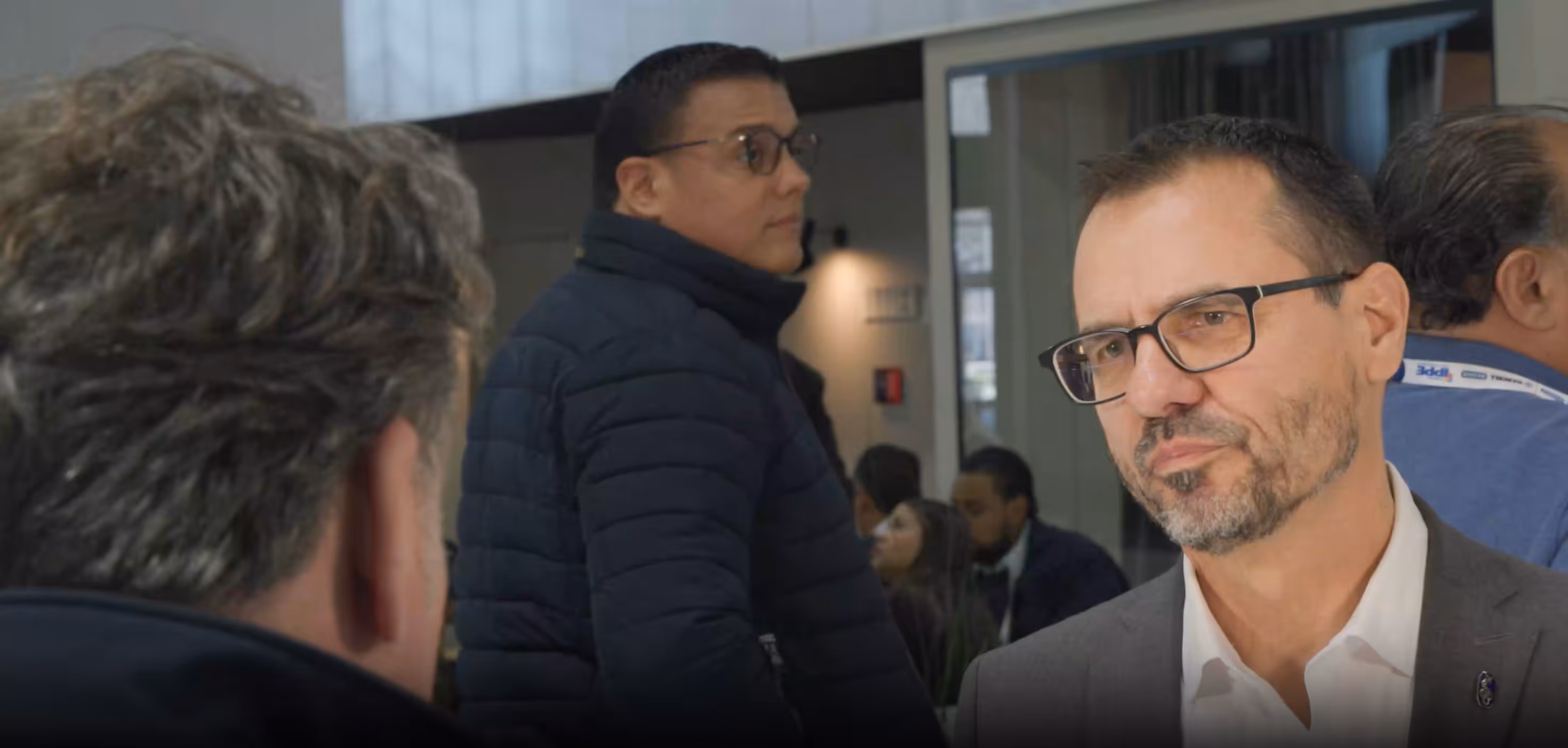 Middle-aged man with glasses and beard conversing with another man, with other people in the background at an indoor event.