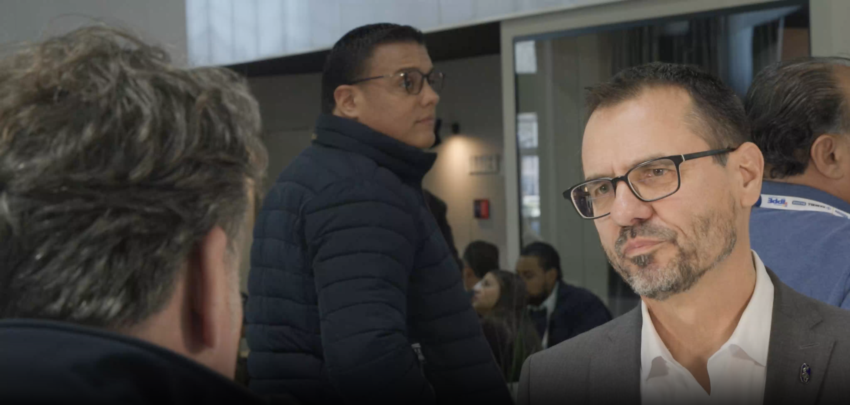 Middle-aged man with glasses and beard conversing with another man, with other people in the background at an indoor event.