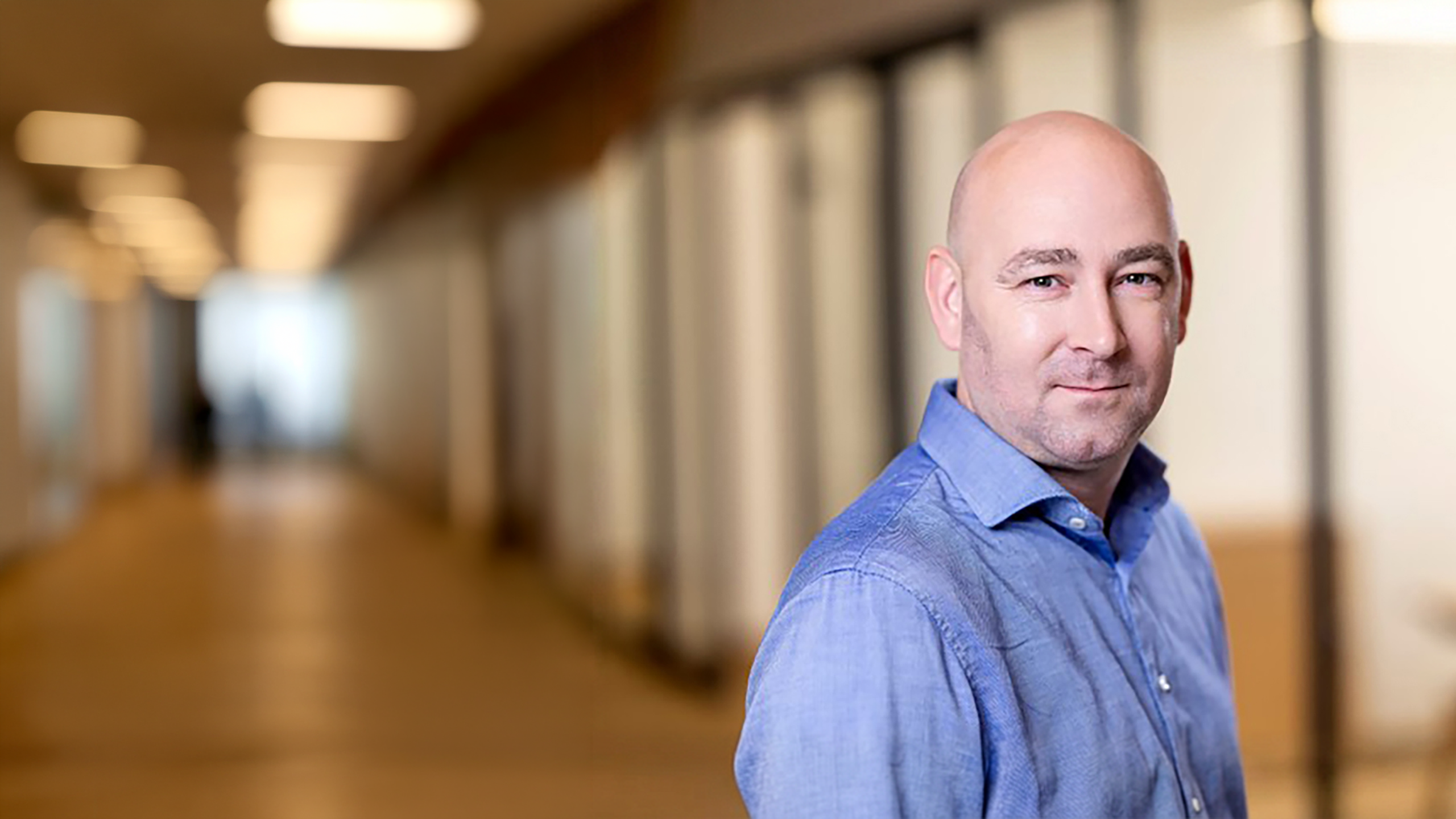 Bald man in a blue button-up shirt standing in a modern office corridor, smiling slightly at the camera.