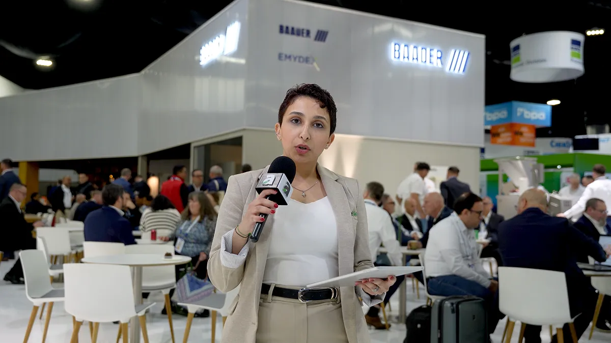 A woman in a beige blazer holding a microphone and a tablet, reporting at a busy conference or trade show with seated attendees in the background.