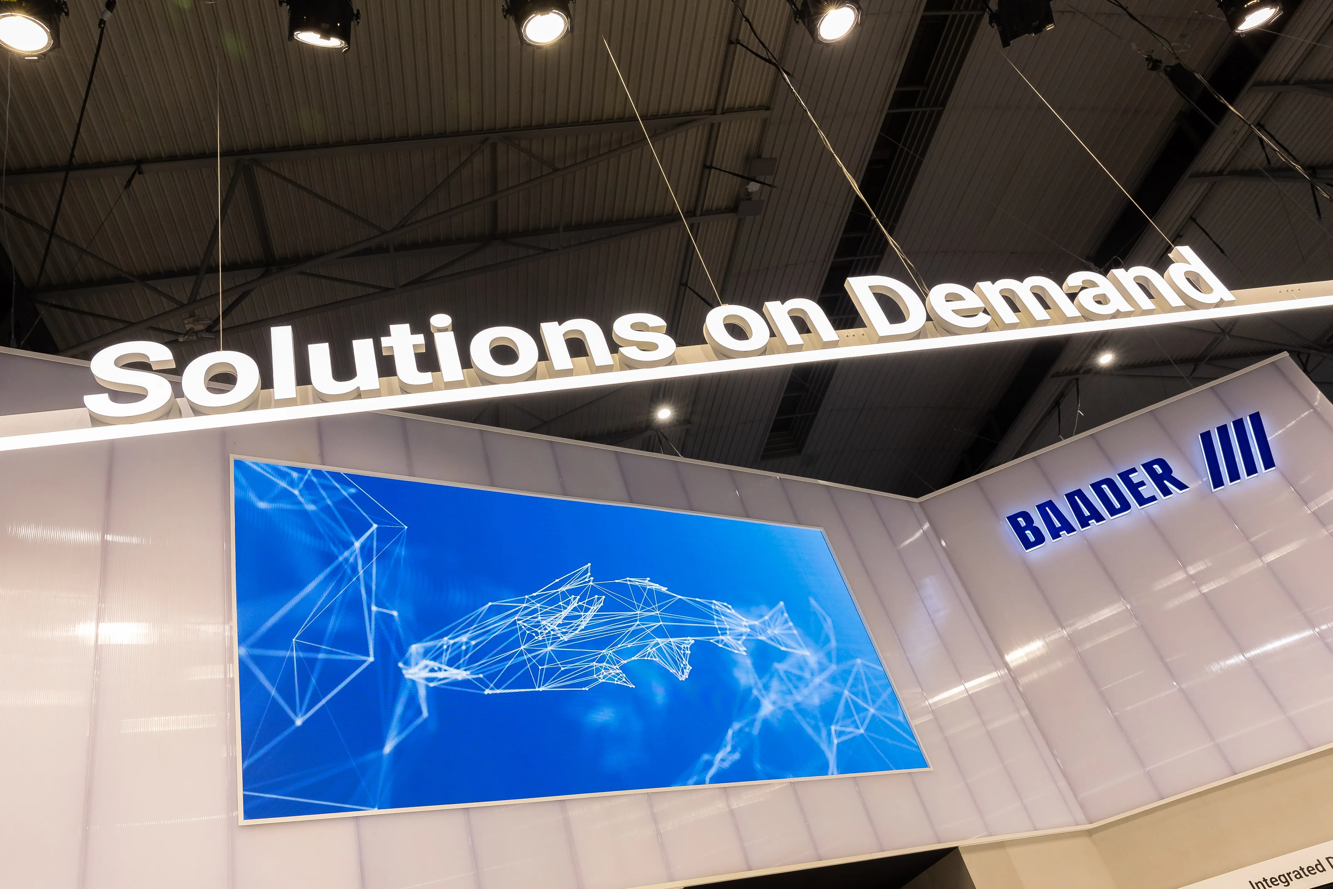 Exhibition booth with illuminated sign 'Solutions on Demand' and a digital display showing a blue wireframe graphic of a fish, Beside the display is the illuminated company name 'BAADER'.