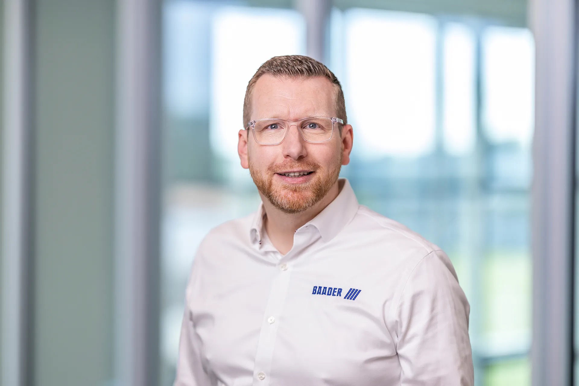 Man with short hair and beard wearing clear glasses and a white shirt with blue 'BAADER' logo smiling in an office setting.