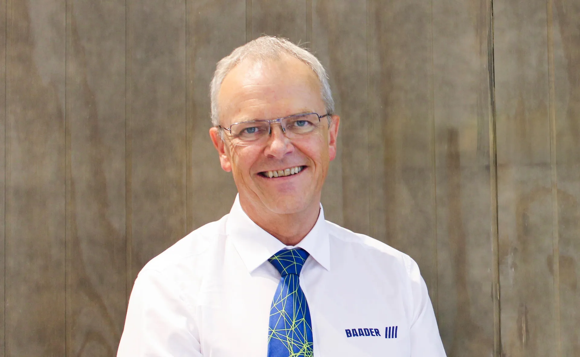 Smiling middle-aged man with gray hair, glasses, white shirt, and blue patterned tie standing in front of a plain, brownish wall.