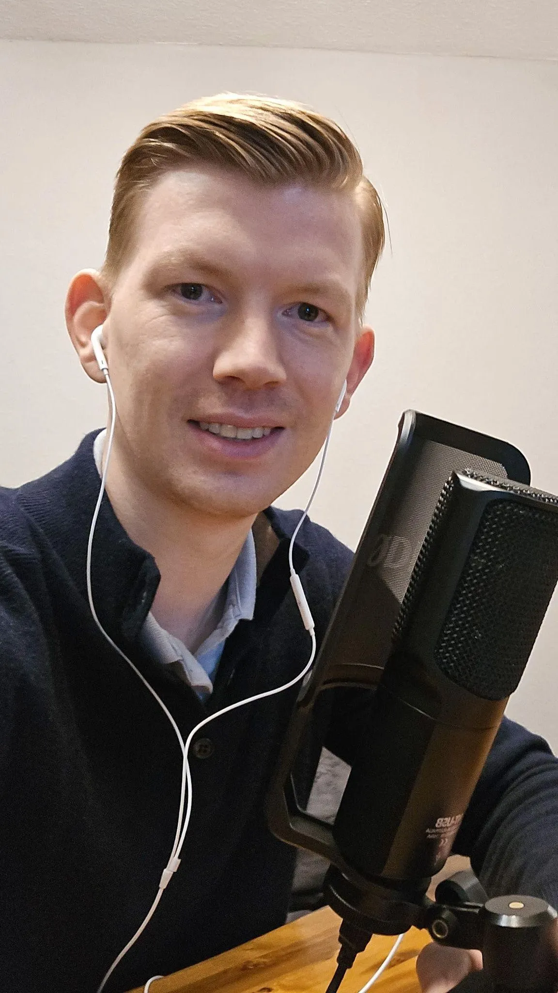 Young man with short blonde hair wearing white earphones, sitting at a wooden table speaking into a black podcast microphone.