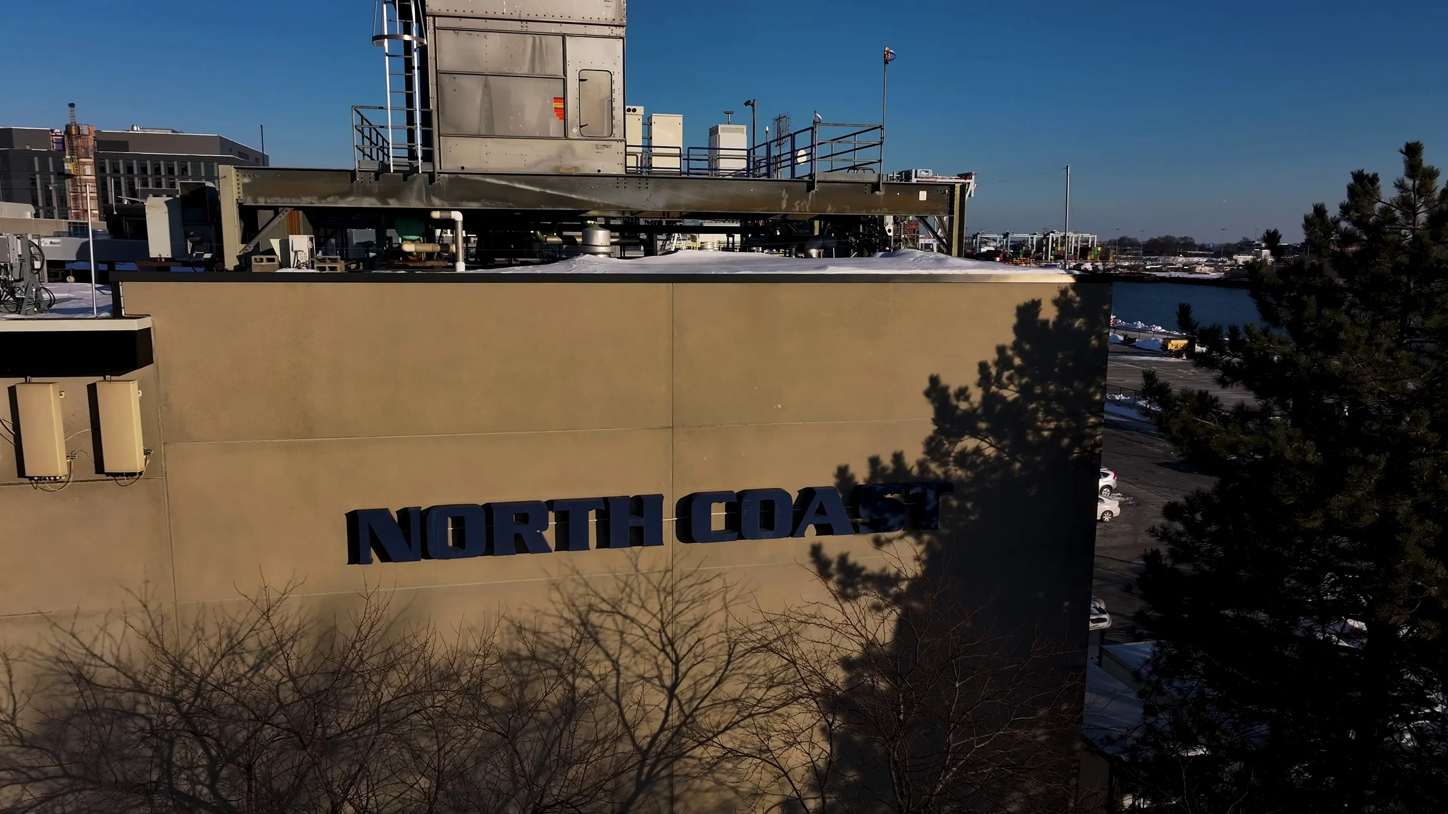 Building wall with large black letters spelling 'NORTH COAST' and shadows of trees on the wall, industrial structures and water in the background.