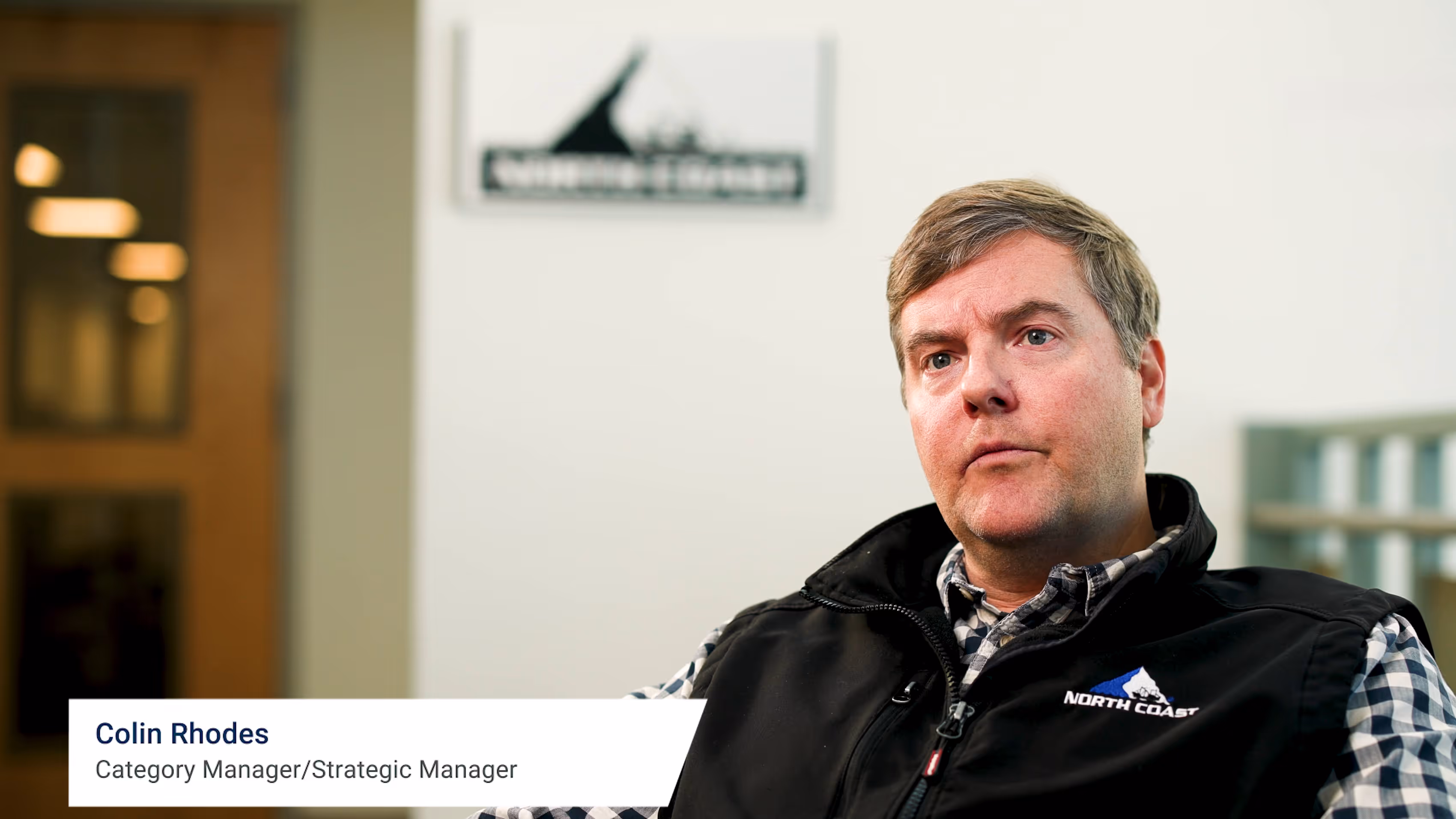 Man wearing a checkered shirt and black North Coast vest, sitting indoors with a serious expression.