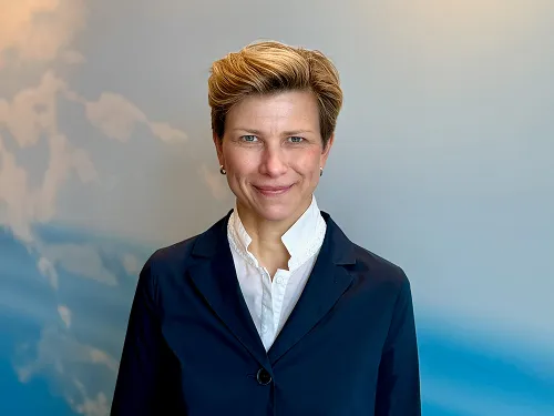 Smiling person with short blonde hair wearing a dark blazer and white collared shirt standing against a blue sky background with clouds.