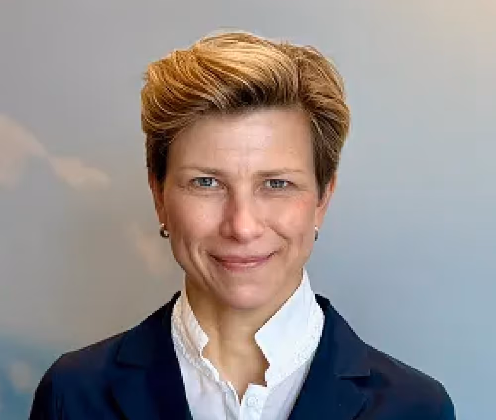Portrait of a person with short blonde hair wearing a dark jacket and white shirt smiling slightly against a gray background.