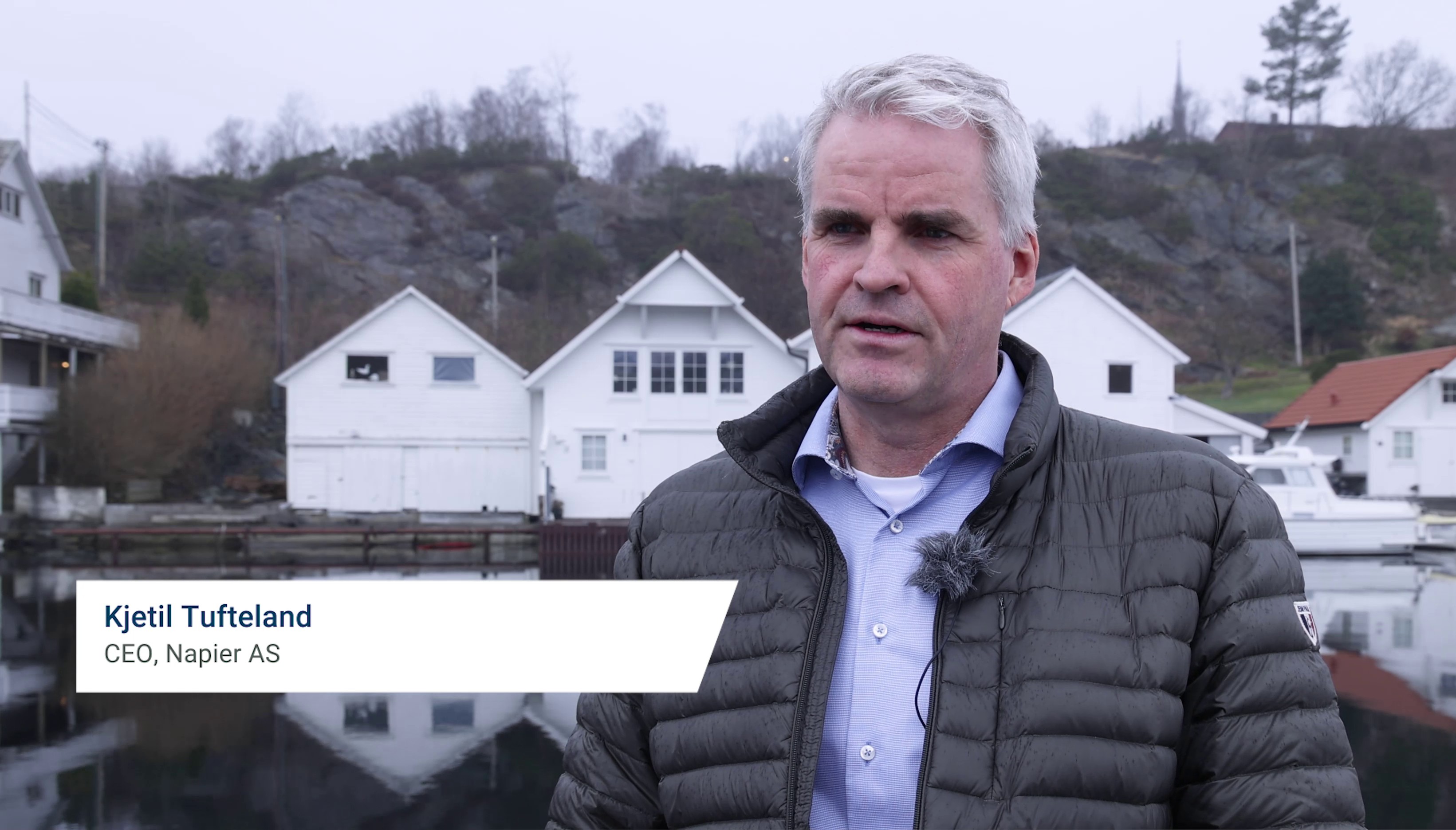 Man with gray hair wearing a black jacket and light blue shirt standing outdoors near a waterfront with white houses and rocky hills in the background.