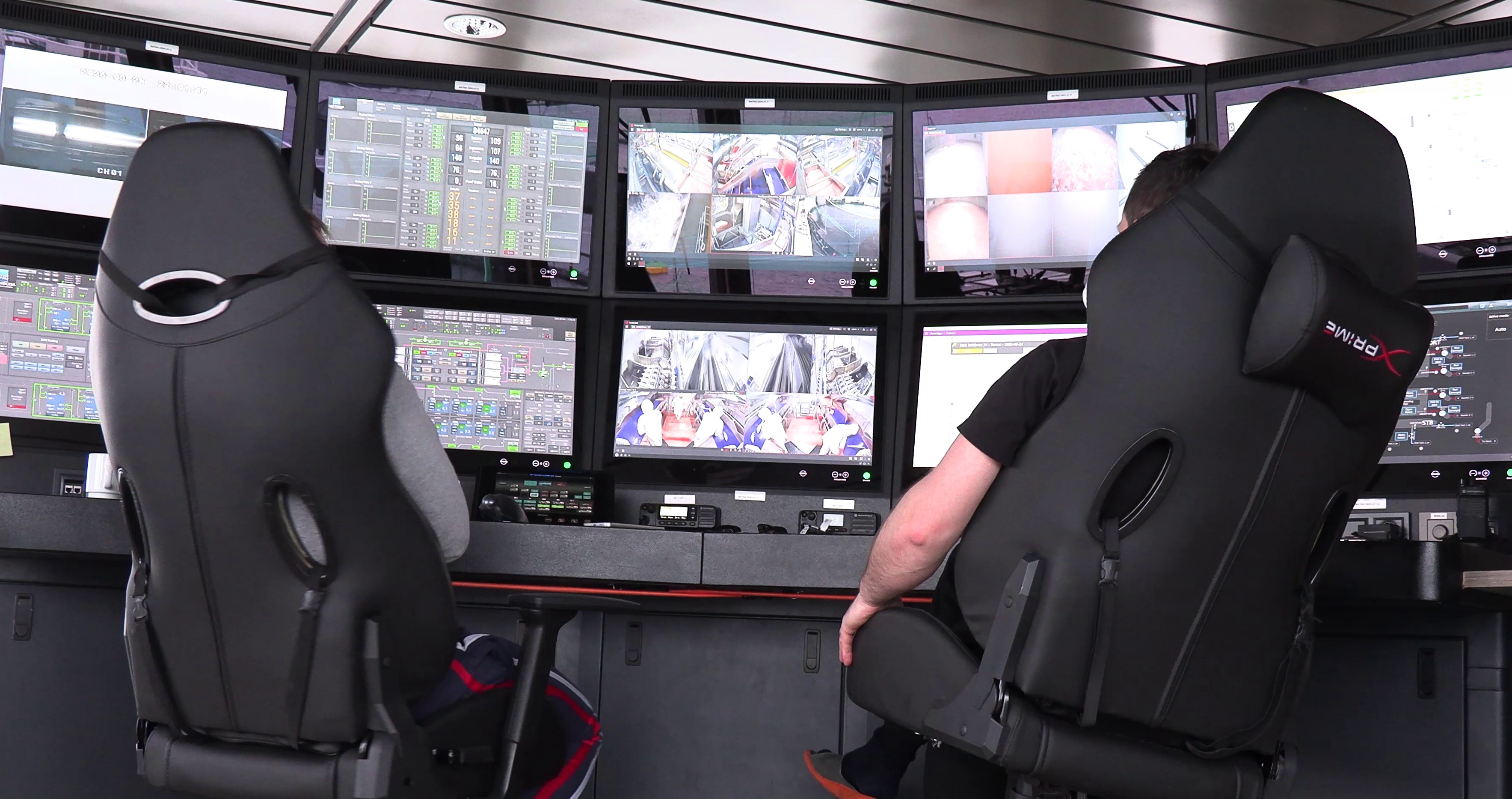 Two people seated in black XPRIME chairs in front of multiple monitors displaying surveillance and control system interfaces.
