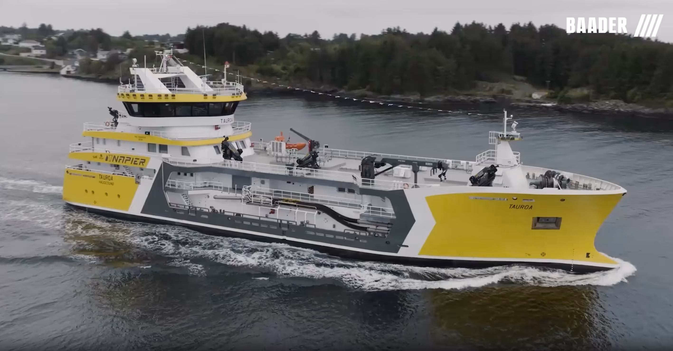 Yellow and white commercial vessel named Tauroa sailing through calm waters near a forested shoreline.