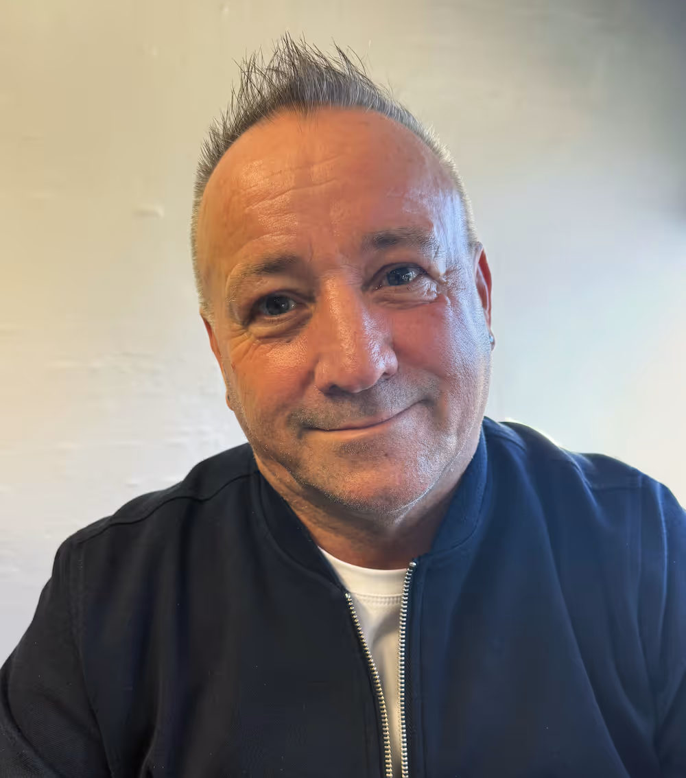 Middle-aged man with short spiked gray hair smiling gently, wearing a black zip-up jacket over a white shirt against a plain light background.