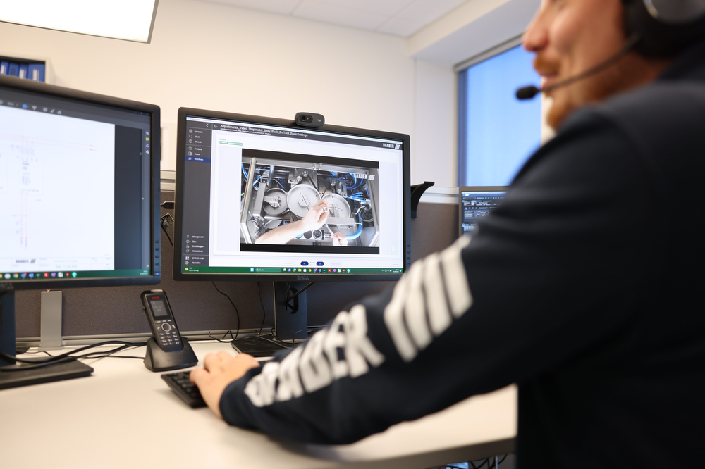 Person wearing headset working at desk with two computer monitors showing technical diagrams and machinery photos.