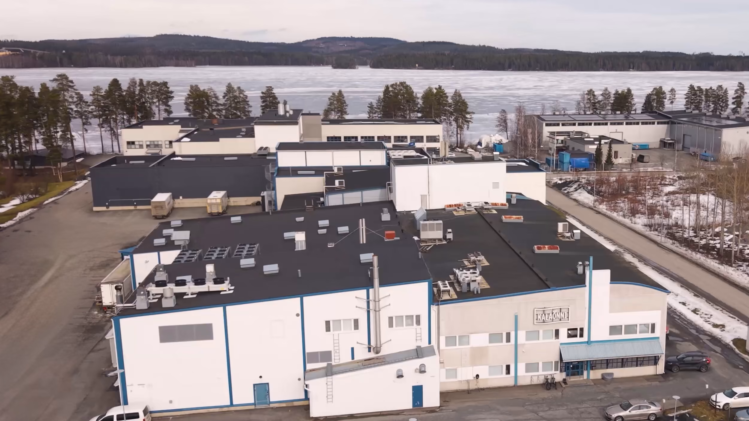 Industrial building complex near a frozen lake with sparse snow and surrounding trees.