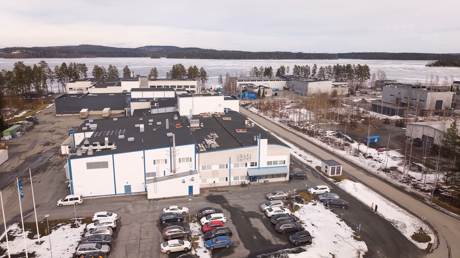 Aerial view of a white and blue industrial building with a parking lot covered in patches of snow near a frozen lake.