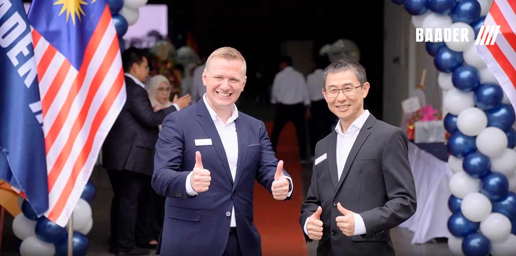 Two businessmen in suits smiling and giving thumbs up between Malaysian flags and blue-white balloon arch.