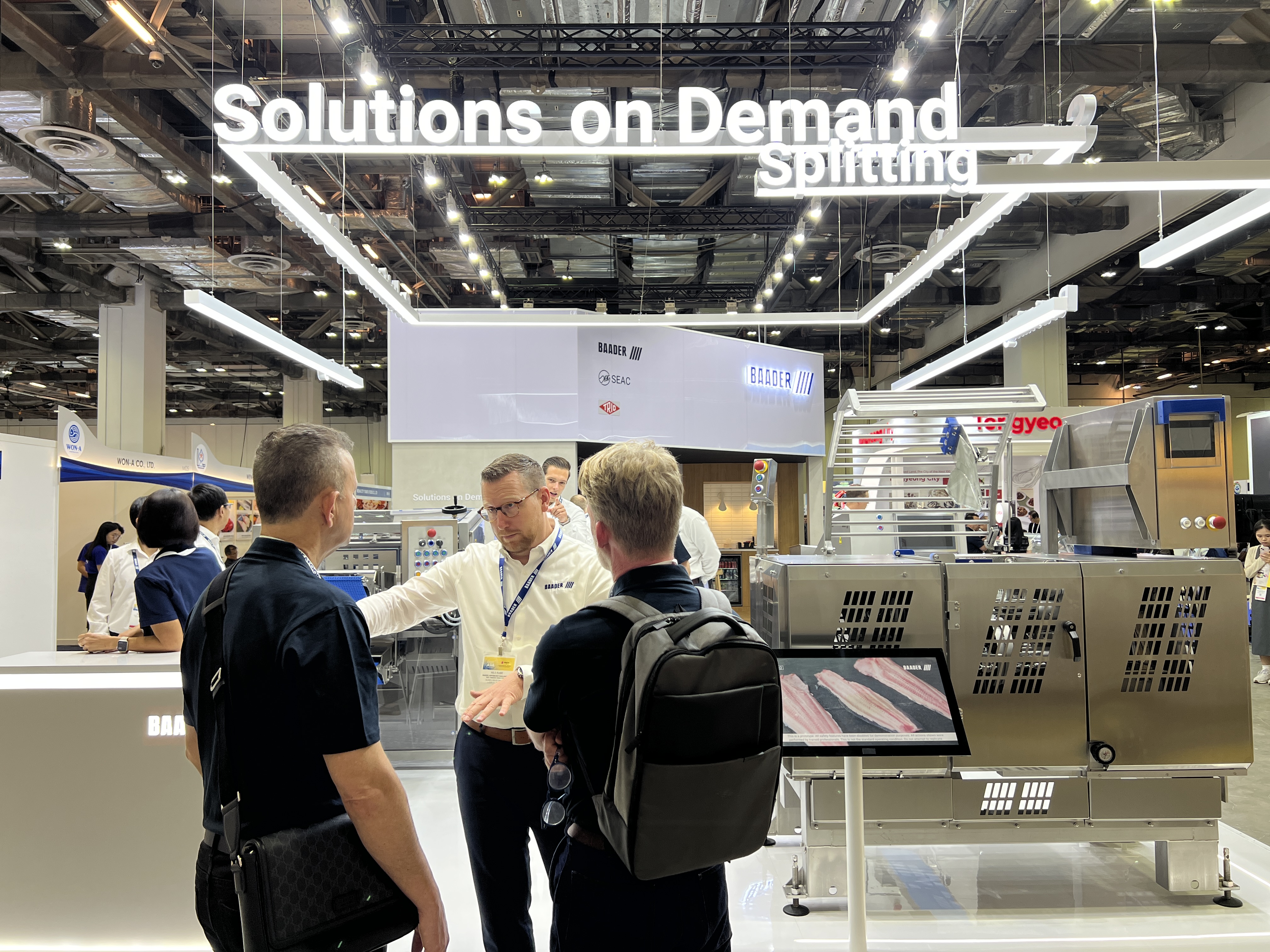 Man in white shirt explaining machinery to two men in black at an industrial trade show booth.