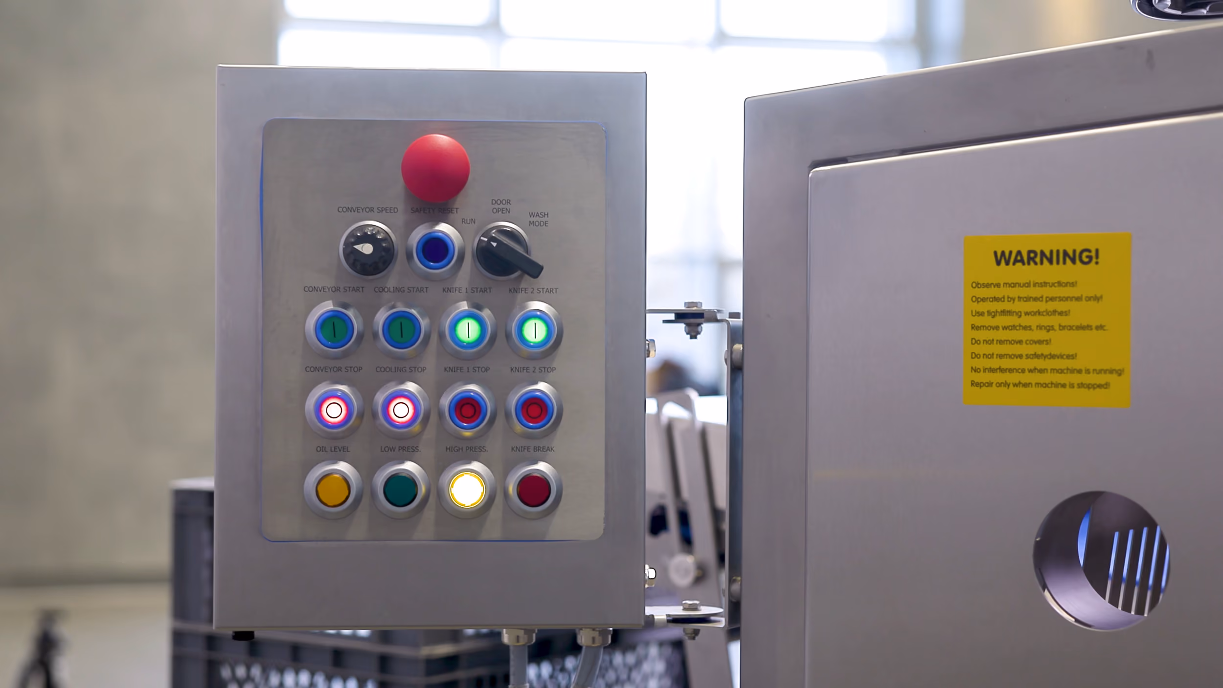 Industrial control panel with multiple colored buttons and switches labeled for conveyor speed, cooling, knife start and stop, oil level, and pressure, mounted on a metallic machine with a yellow safety warning label.