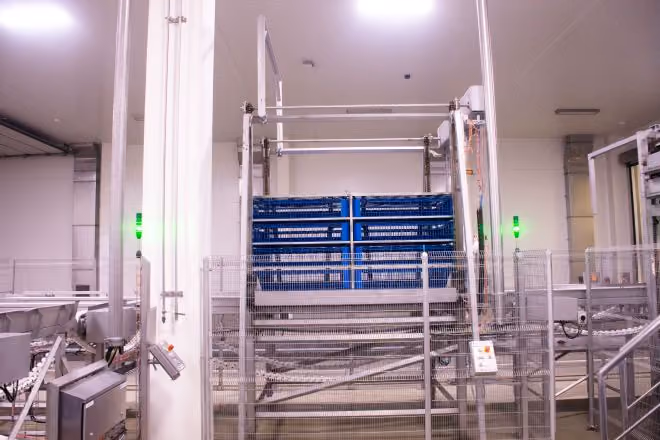 Industrial machinery with blue crates stacked in a white, well-lit factory room surrounded by metal fencing.
