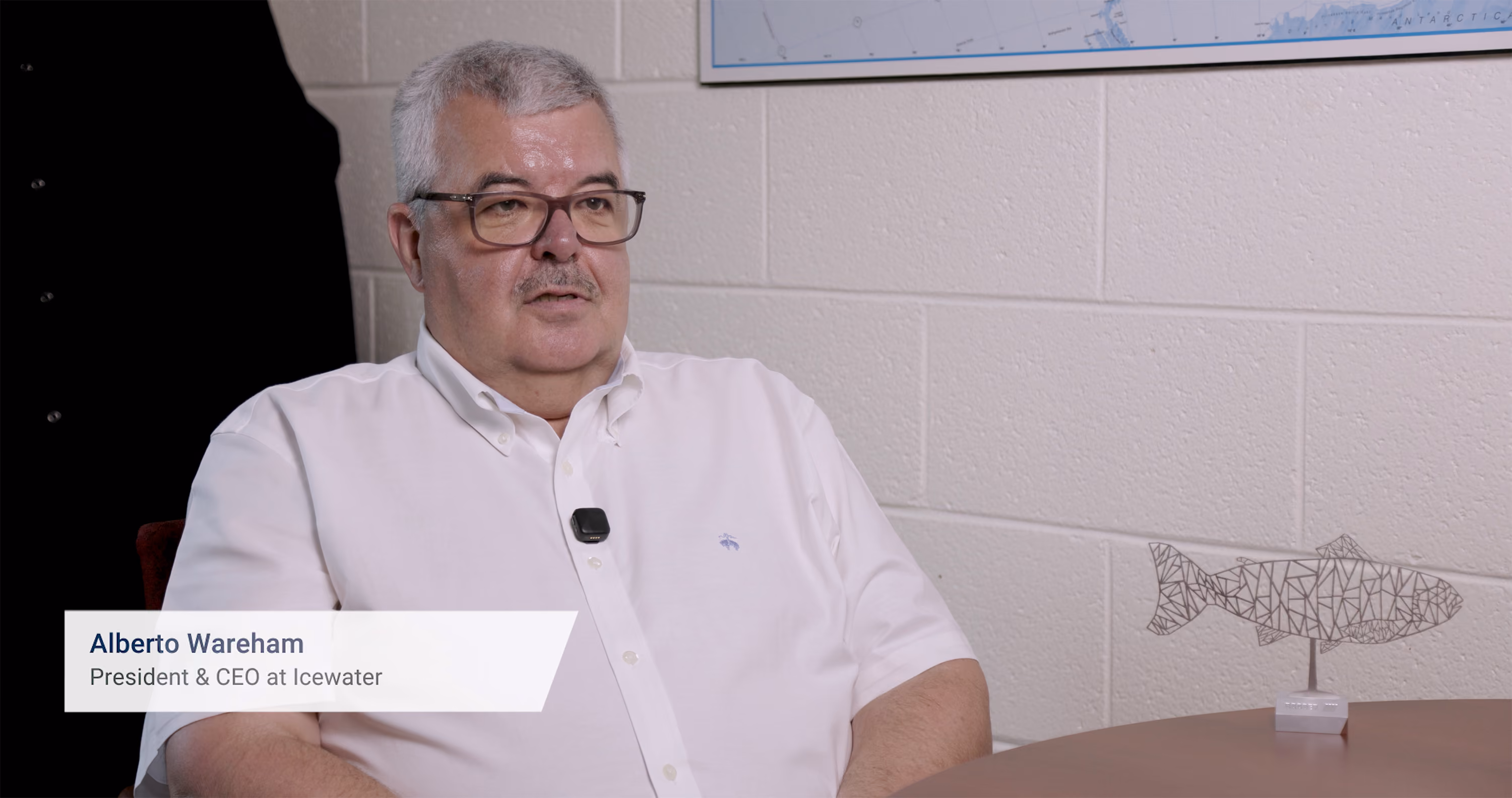 Alberto Wareham, a man with short gray hair and glasses, wearing a white button-up shirt, seated indoors with a microphone clipped to his shirt, next to a table with a geometric metal fish sculpture and a map on the wall behind.