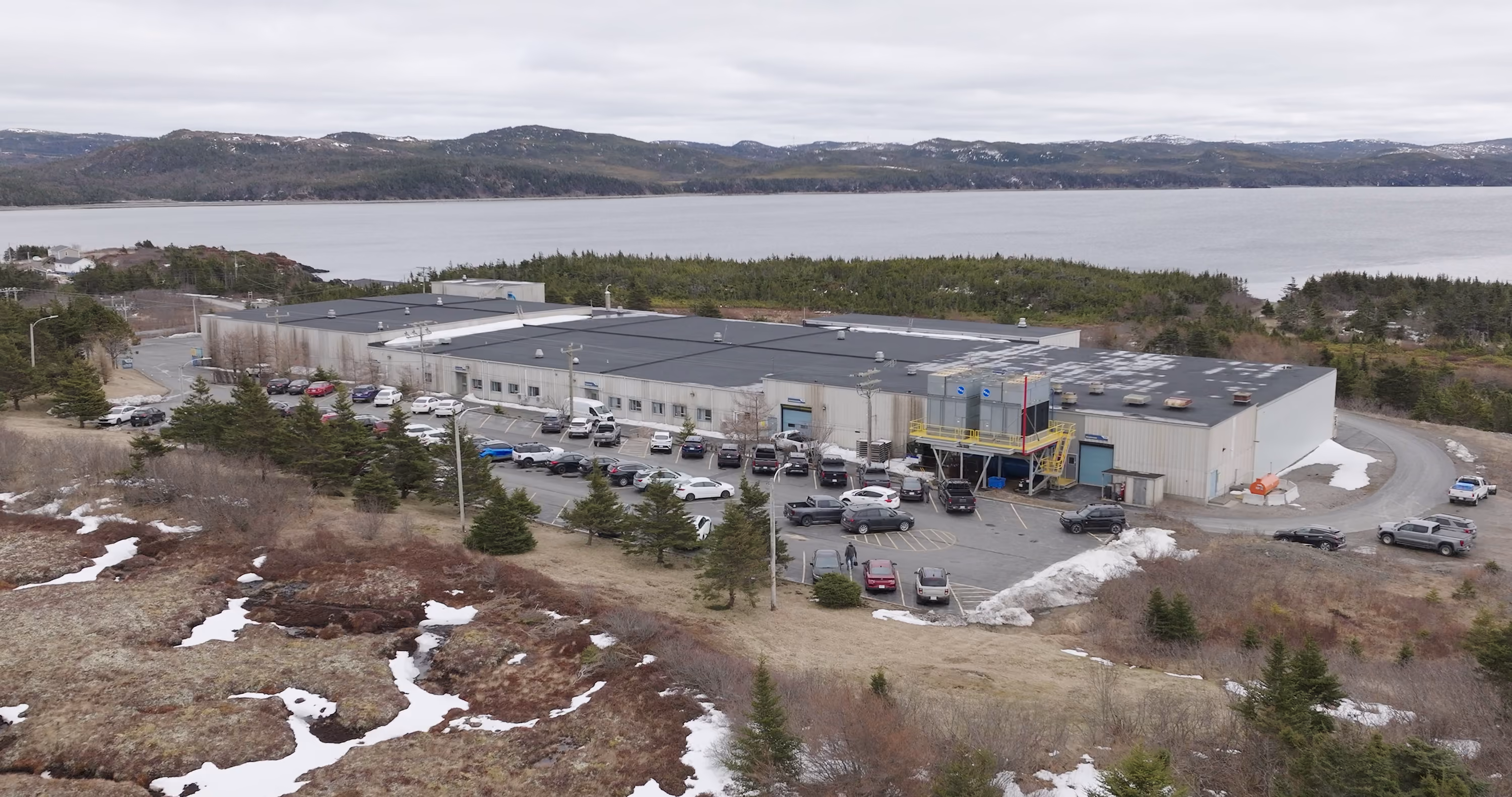 A large rectangular industrial building with a flat black roof surrounded by parked cars, set near a body of water with forested hills in the background and patches of snow on the ground.