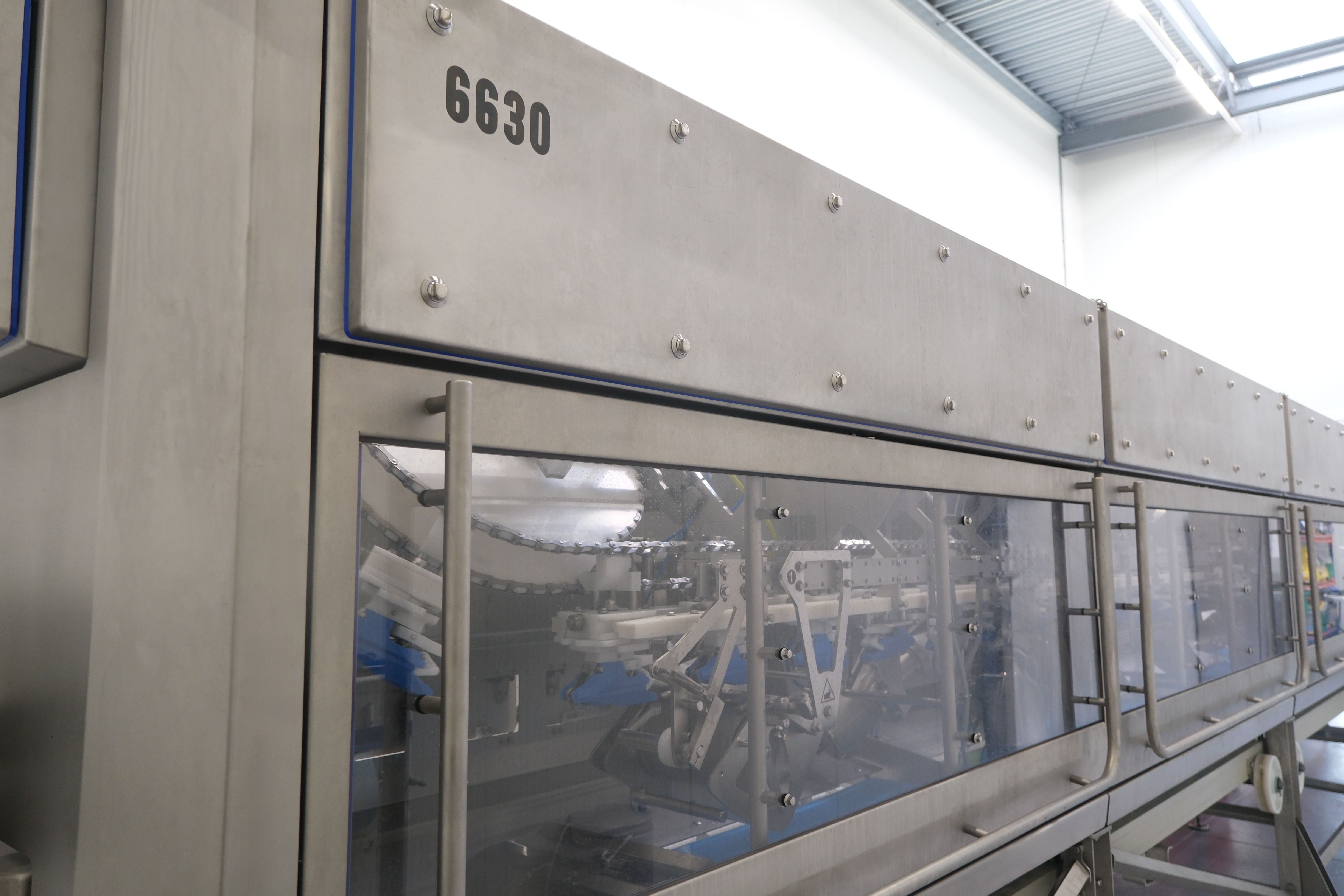 Large industrial stainless steel machine with glass panel showing internal mechanical components, labeled 6630, in a bright factory setting with metal roof and lighting above.