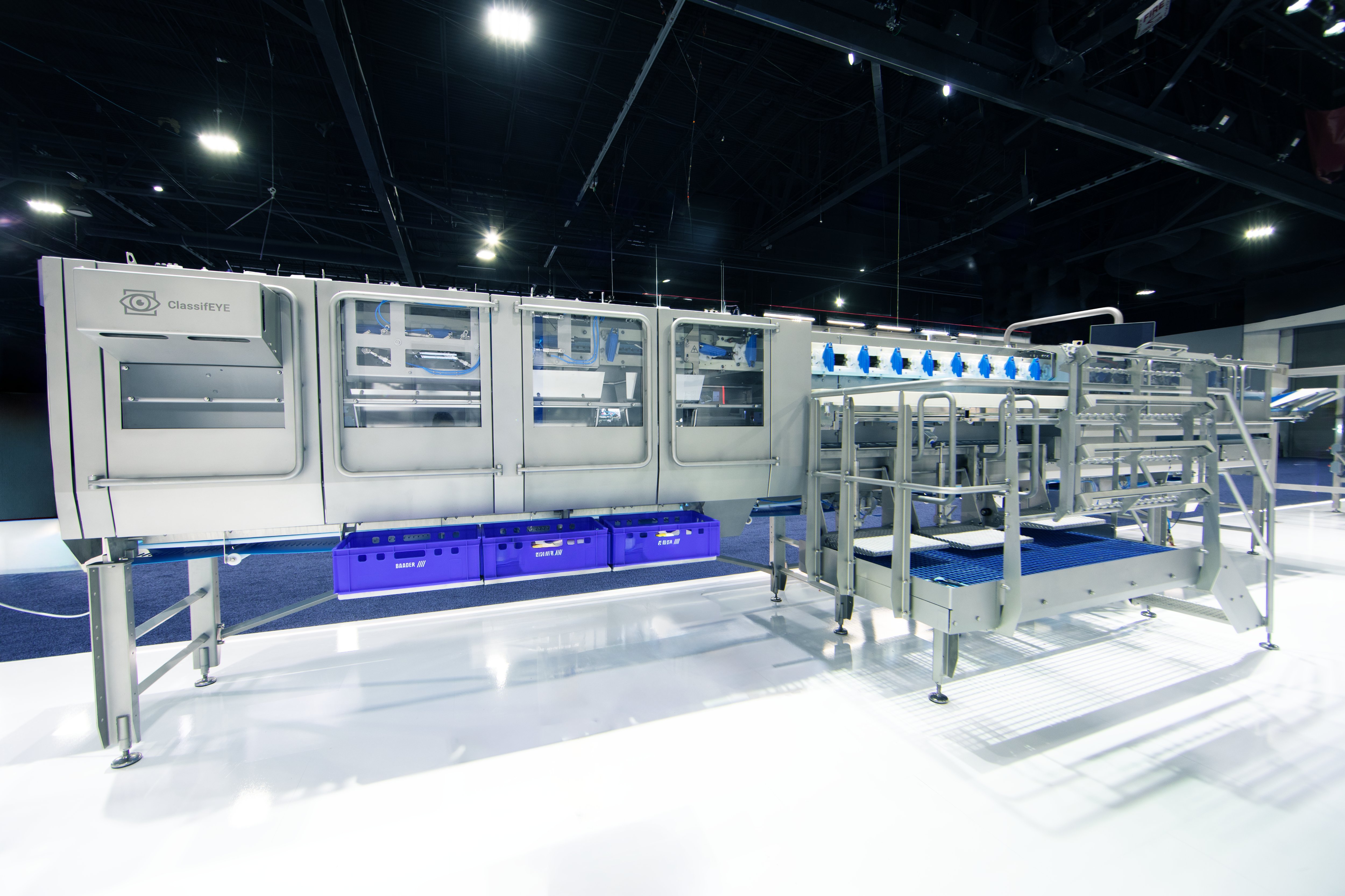 Large industrial machine labeled ClassifEYE with enclosed sections, conveyor belts, blue crates underneath, and metal railings and stairs on a shiny white floor in a spacious indoor facility.