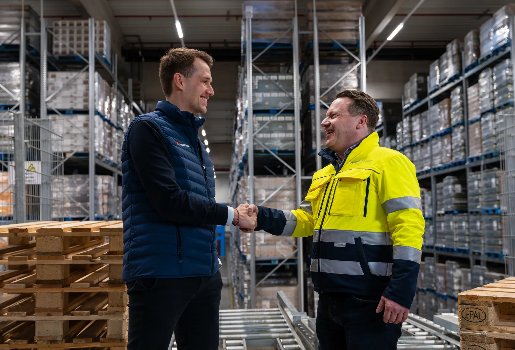 Dr. Philipp Hüning, Co-Founder & CEO Logistikbude (links), Jens Kleiner, COO der Nagel-Group geben sich die Hand und lachen sich an, in einem Palettenlager