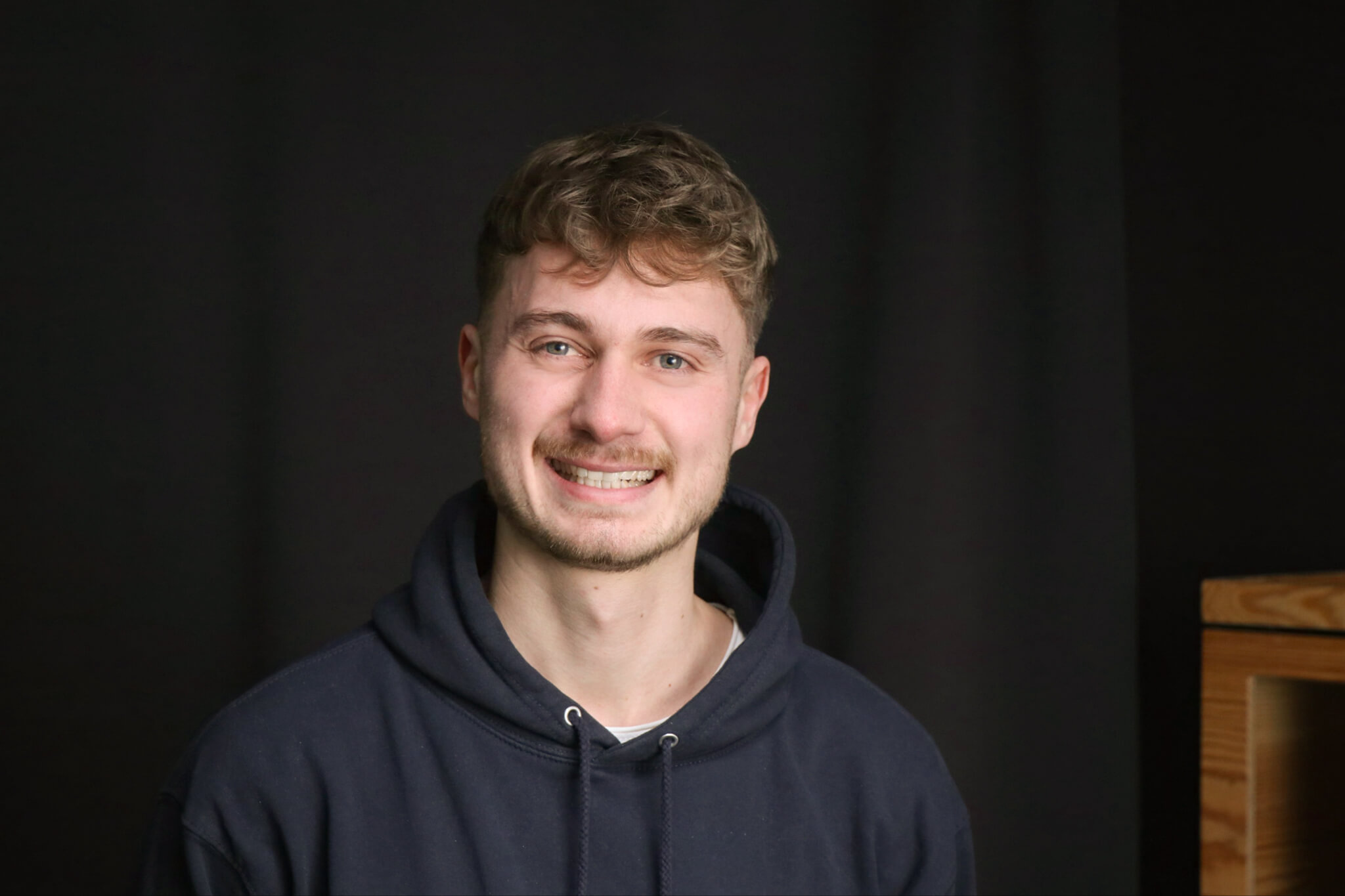 A young man with curly hair and a dark blue hoodie smiles friendly into the camera. The background is dark and simple