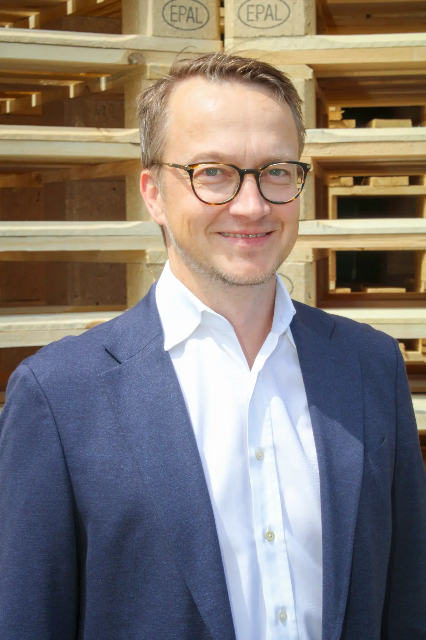 A smiling man with glasses and a jacket stands in front of a stack of EPAL Euro pallets. He is wearing a white shirt and looking directly into the camera.