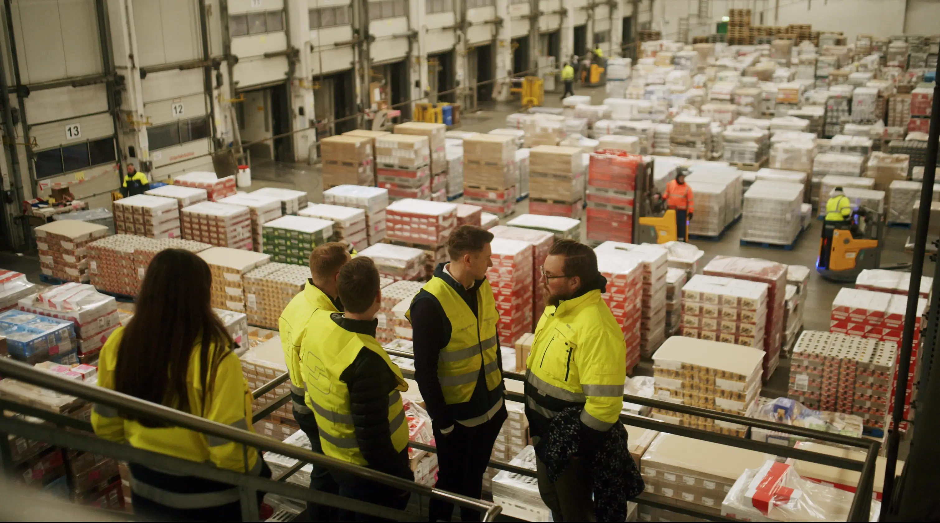 Mehrere Personen in gelben Warnwesten stehen in einer großen Logistikhalle und blicken auf gestapelte Paletten mit Waren. Im Hintergrund sind Mitarbeitende auf Flurförderfahrzeugen unterwegs.