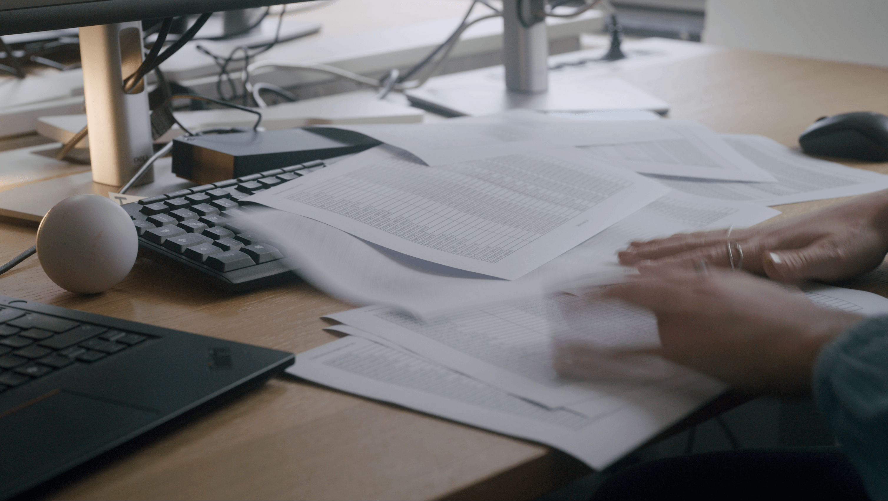 Messy desk with lots of printed spreadsheets being searched through and sorted by one person. Next to it is a laptop, keyboard and monitor.